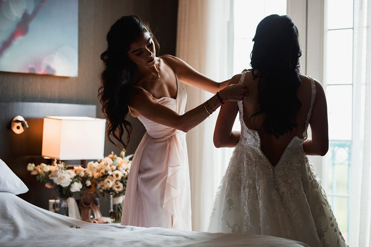 a bride and her sister in elegant white dresses help each other get ready in a sunlit room with flowers and soft decor.
