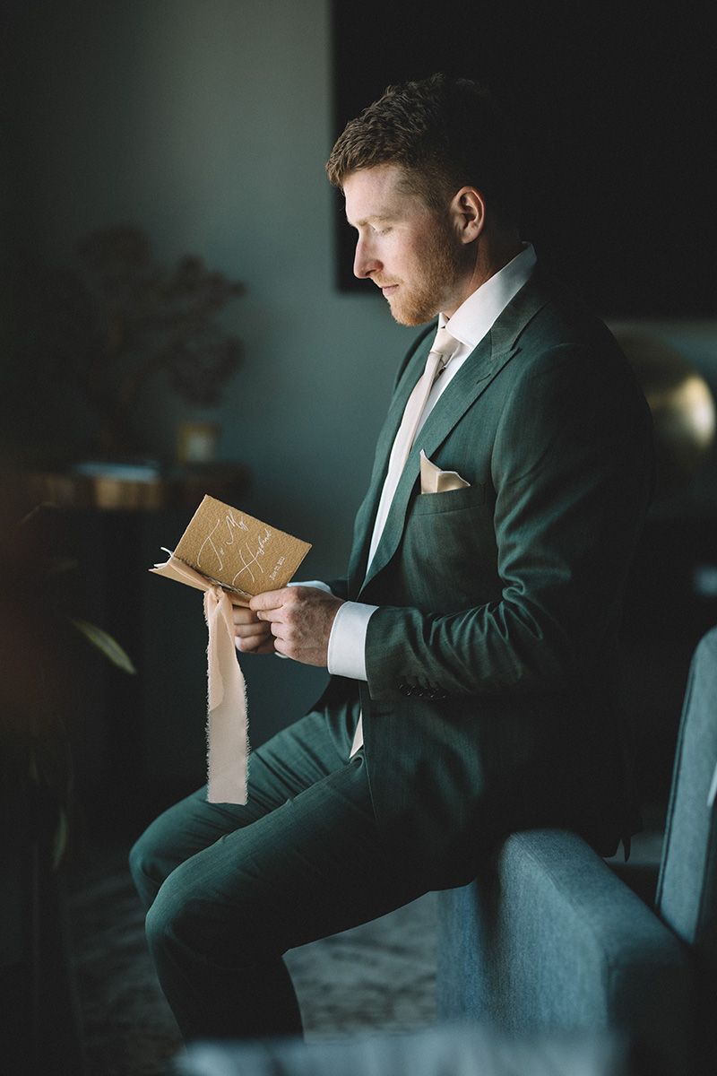 A groom in a dark green suit sits in a chair reading a letter in a moody, atmospheric setting.