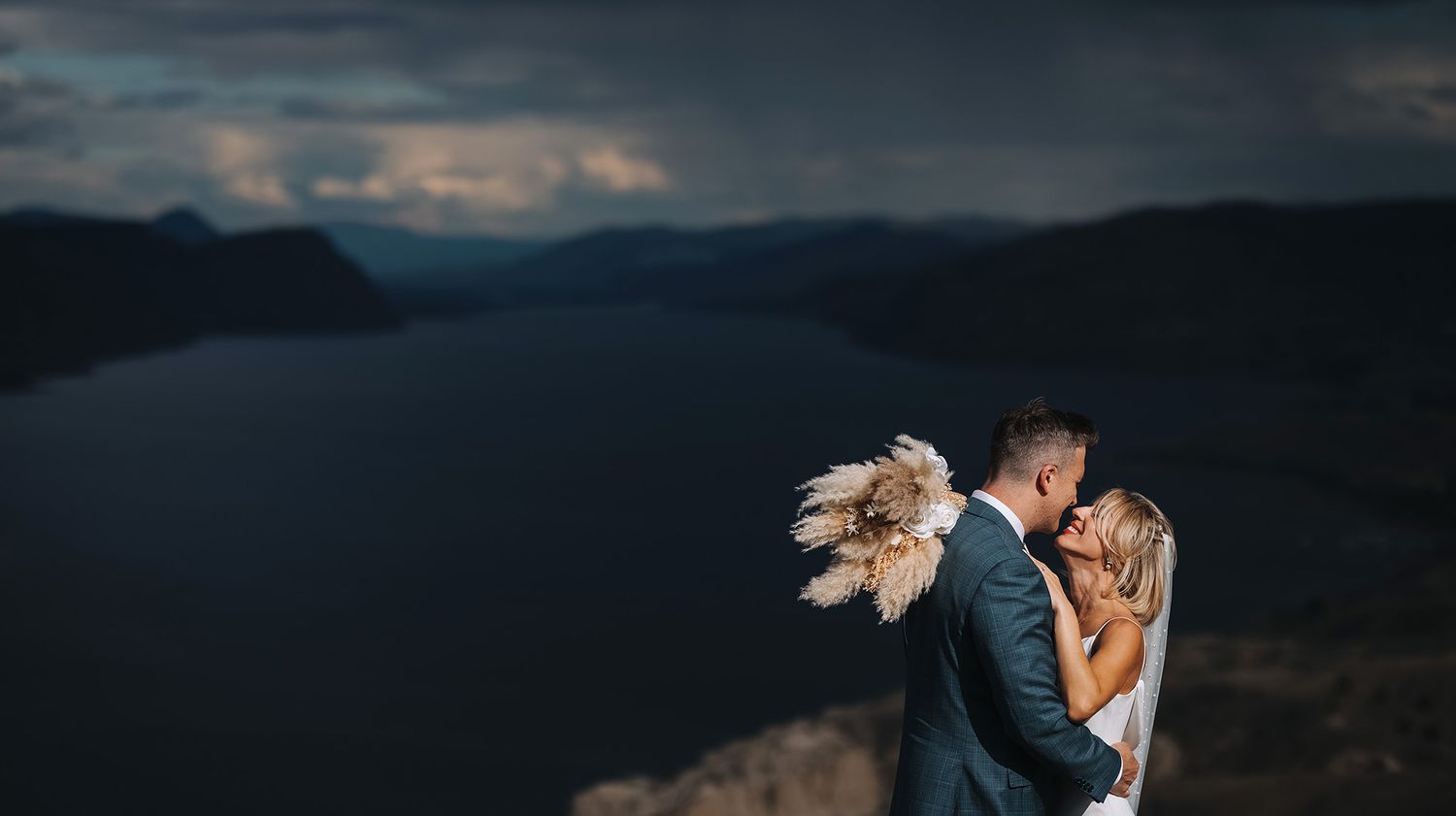 A couple embraces on a mountain overlook at dusk with dramatic storm clouds, lake and mountains in the background.