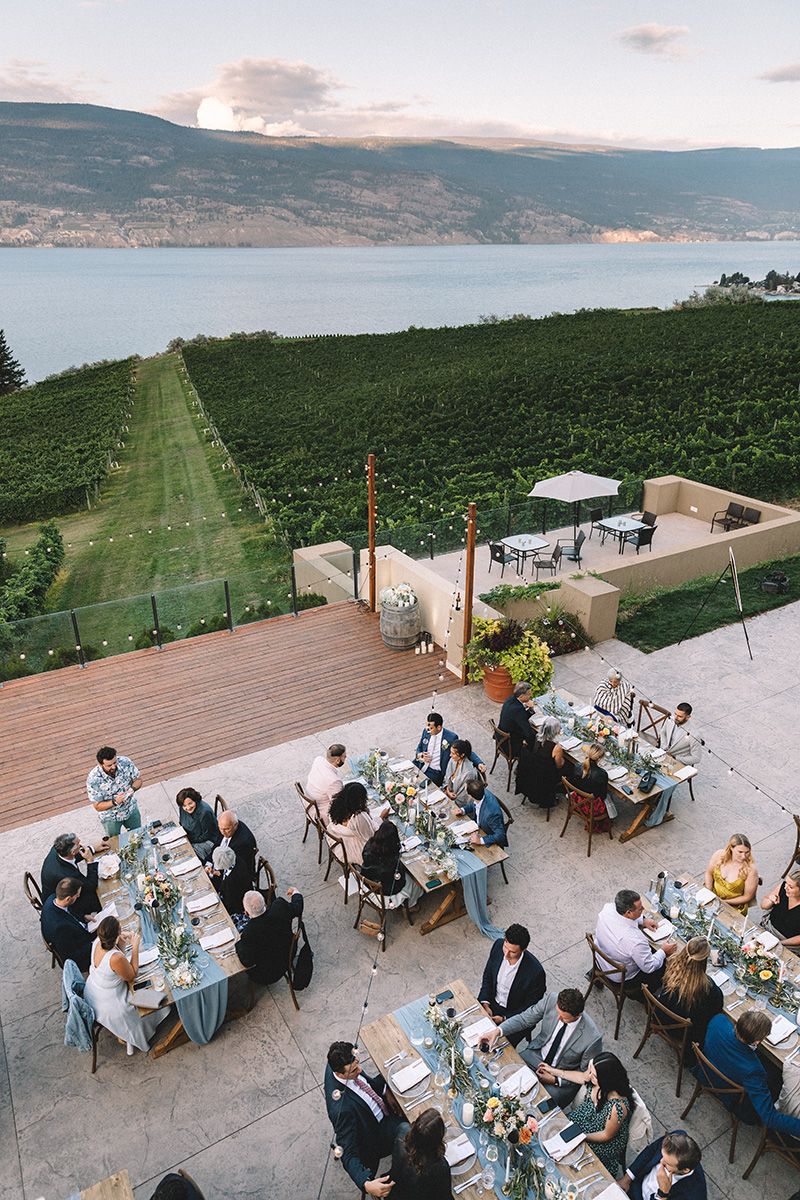 Aerial view of an outdoor wedding reception on a terrace overlooking vineyards and a scenic lake at sunset.