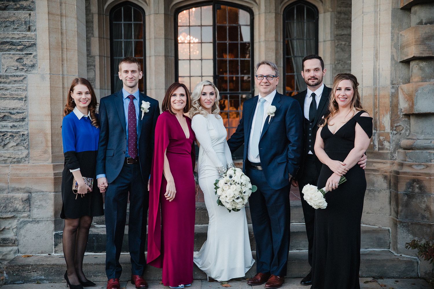 A wedding party poses together in front of a stone church exterior in formal attire.