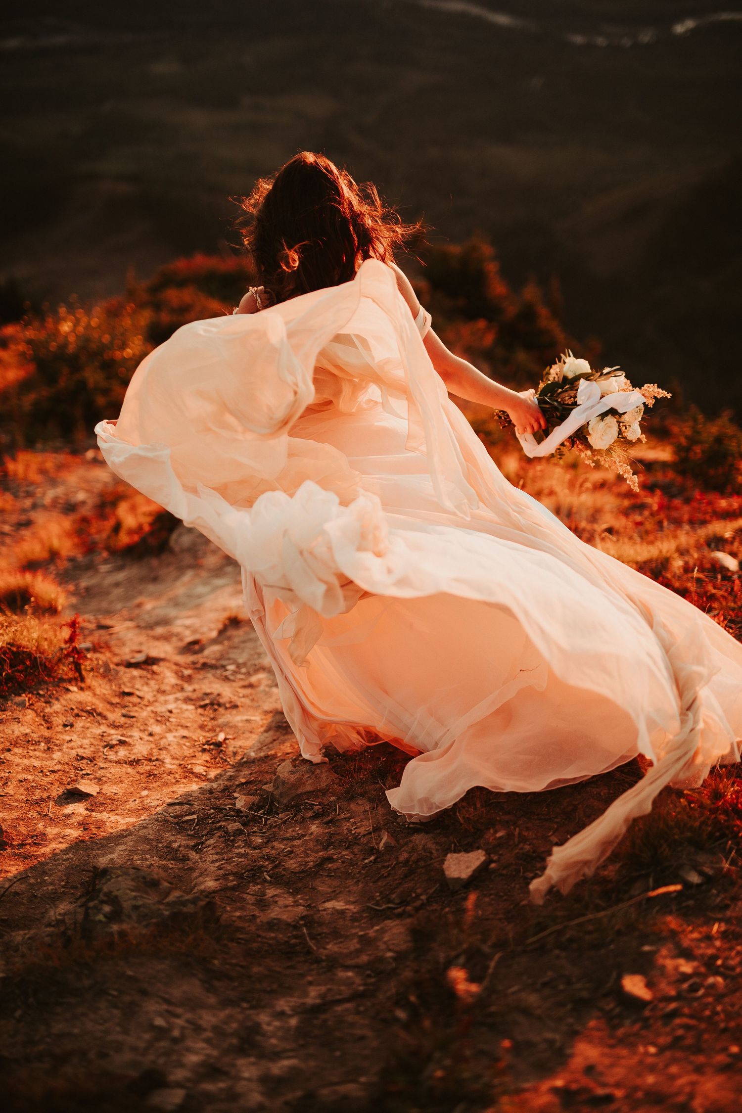 A bride in a flowing wedding gown runs down mountain path with wind billowing her dress.