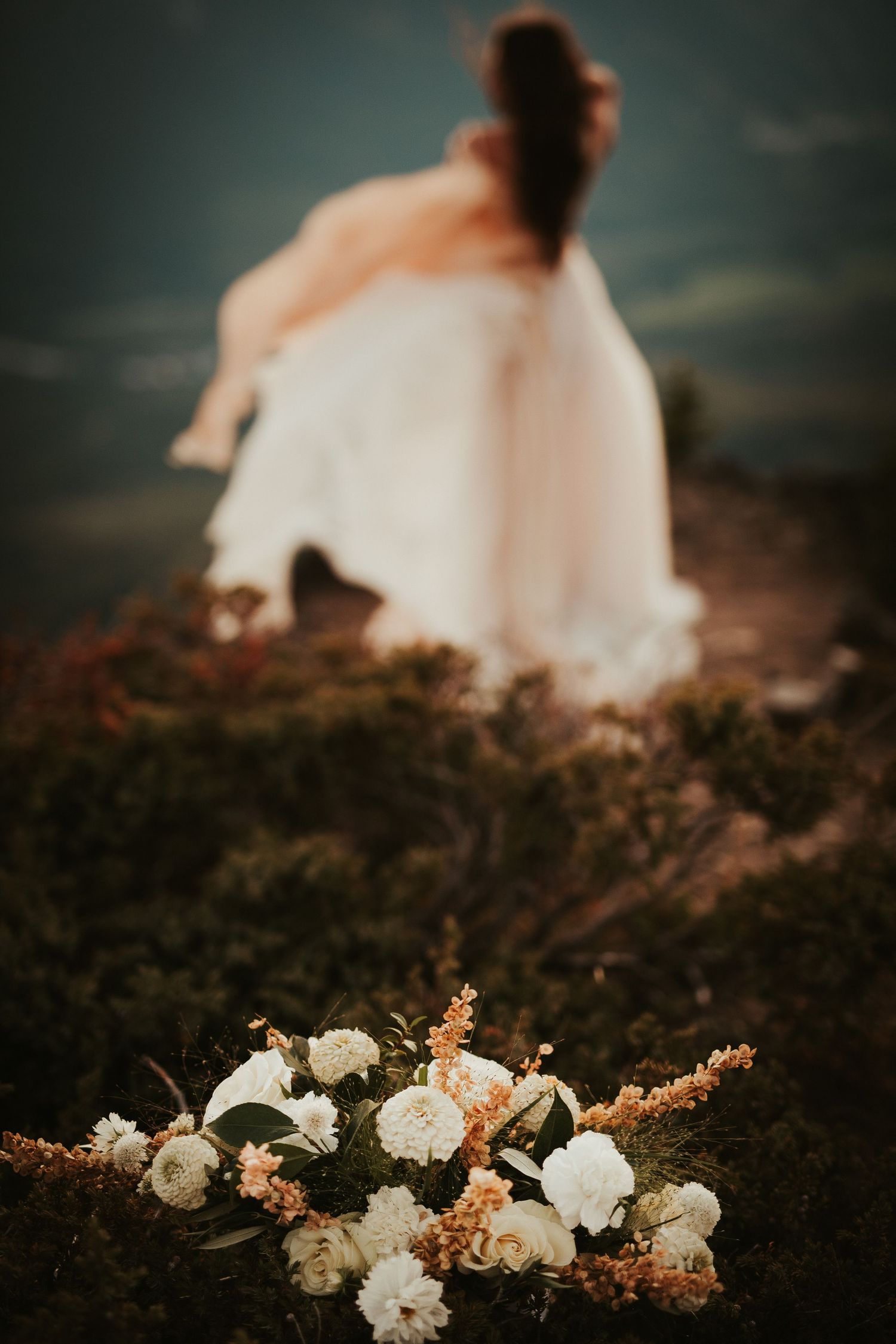 White flowers lay scattered on dark ground with ethereal white dress details visible in a moody outdoor setting.