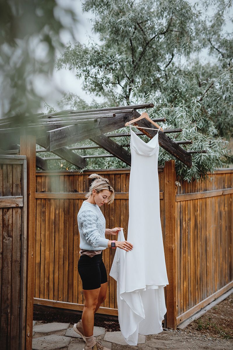 A white wedding dress hangs from a tree branch against a wooden fence while a bride admires it.