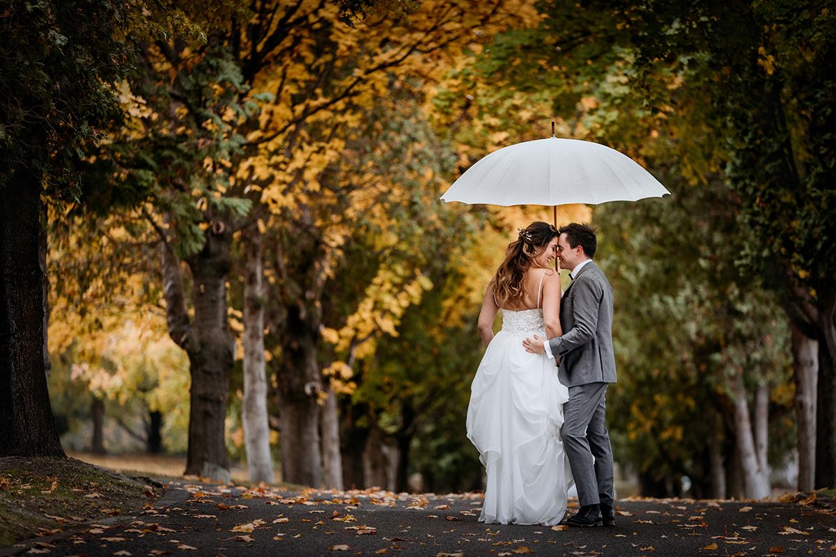 A couple shares a romantic moment under a white umbrella in an autumn park with golden foliage.