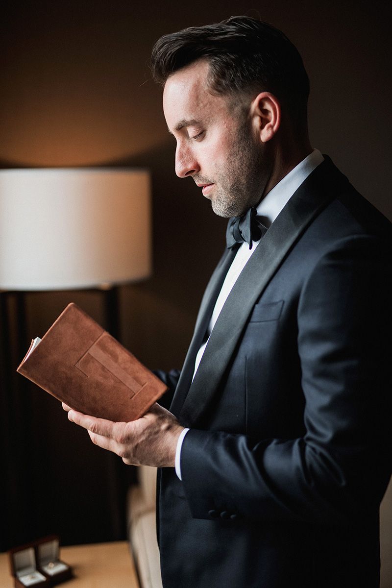 A groom in a black tuxedo and bow tie reads from a brown leather book in moody lighting next to a table lamp.