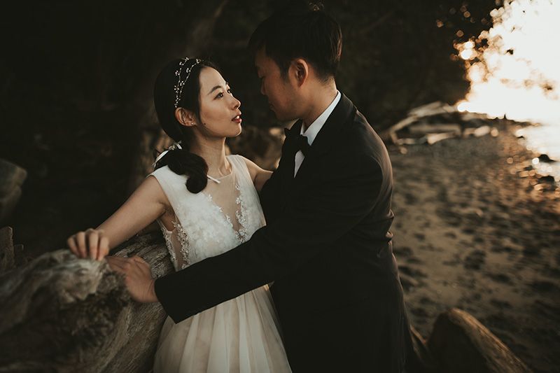 A couple in wedding attire embraces during golden hour at sunset on a scenic beach.