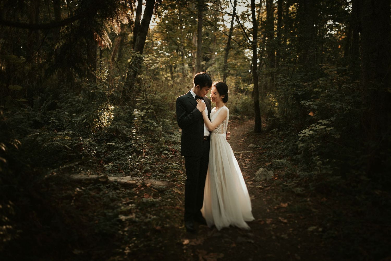 A romantic couple in wedding attire poses together in a serene wooded forest setting at dusk.