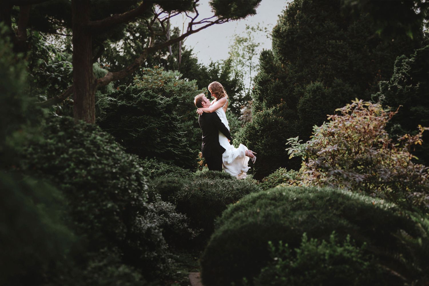A groom lifts his bride in a green garden pathway with stone steps winding through dense foliage and flowering plants in a tranquil setting.
