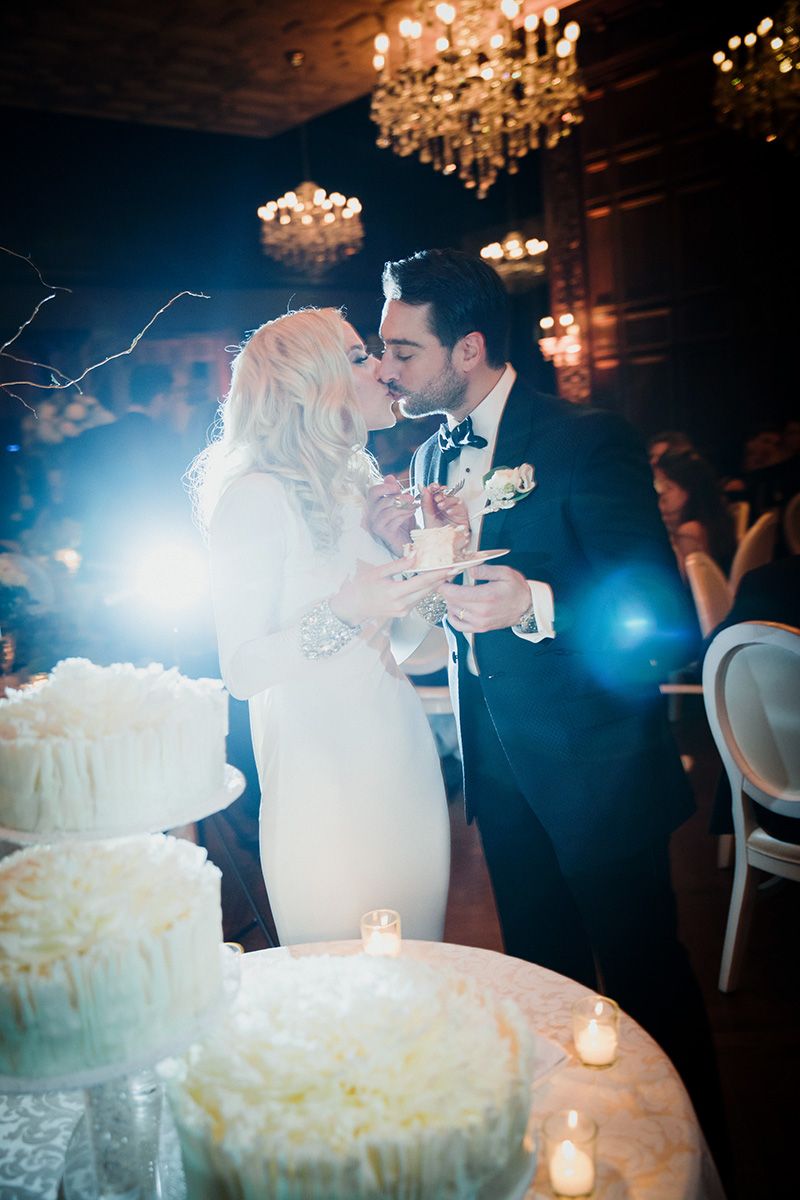 Romantic newlyweds share their first bite of elegant white wedding cake under sparkling crystal chandeliers at reception.