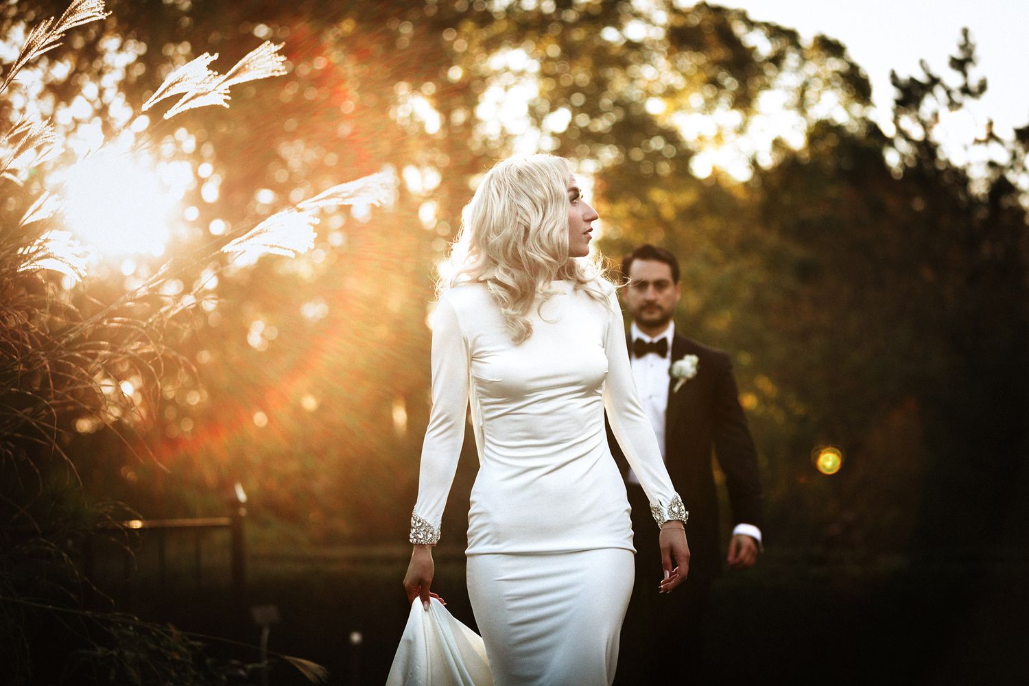 A bride in a white dress walks through a sunlit garden during golden hour as sunbeams filter through the trees.