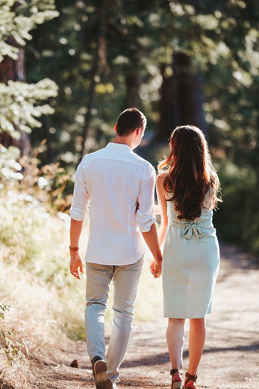 Couple holding hands walking down a sunlit forest path, man in white shirt and woman in light green dress.