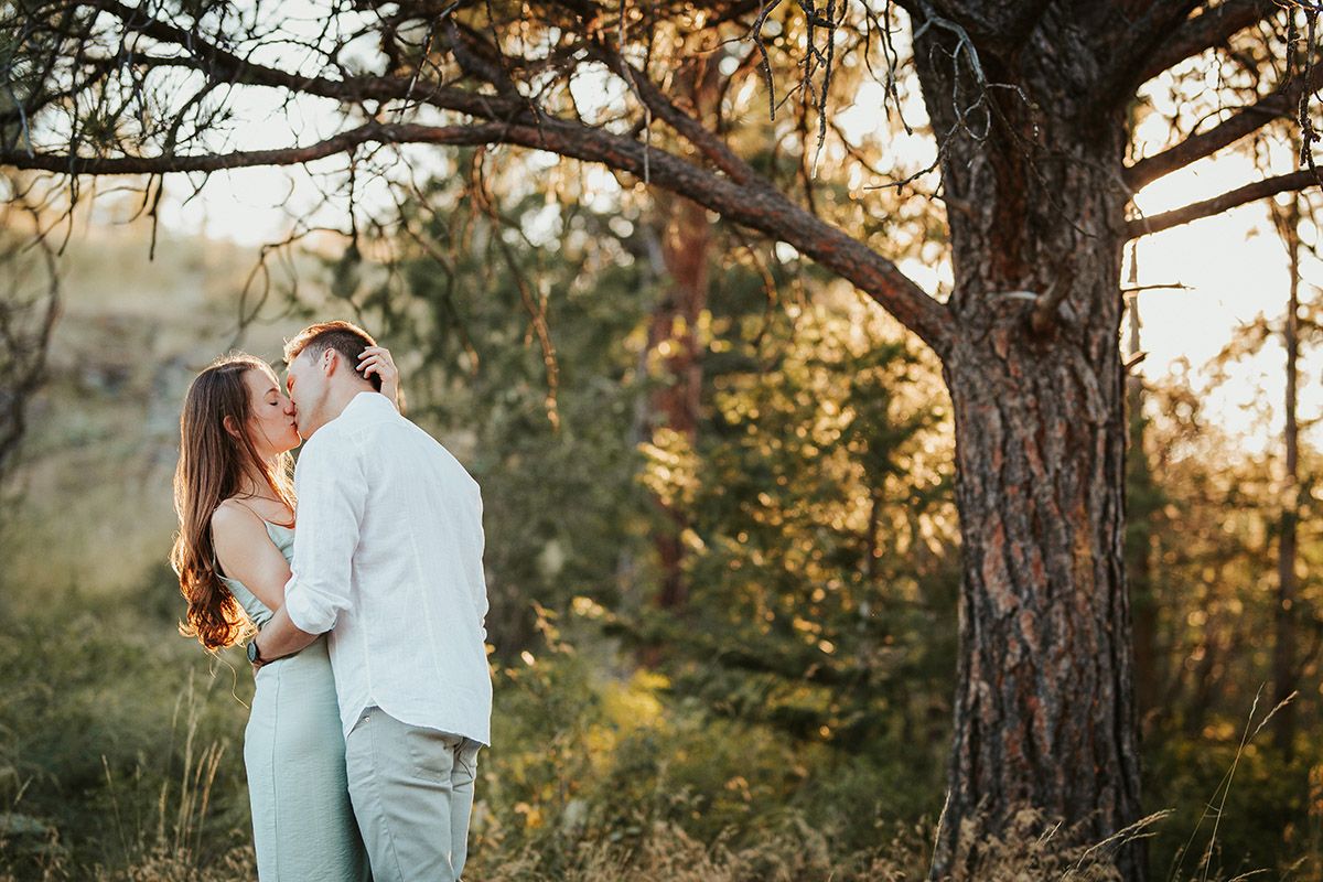Couple sharing a romantic kiss under a large tree in a golden sunlit forest during an outdoor engagement session.