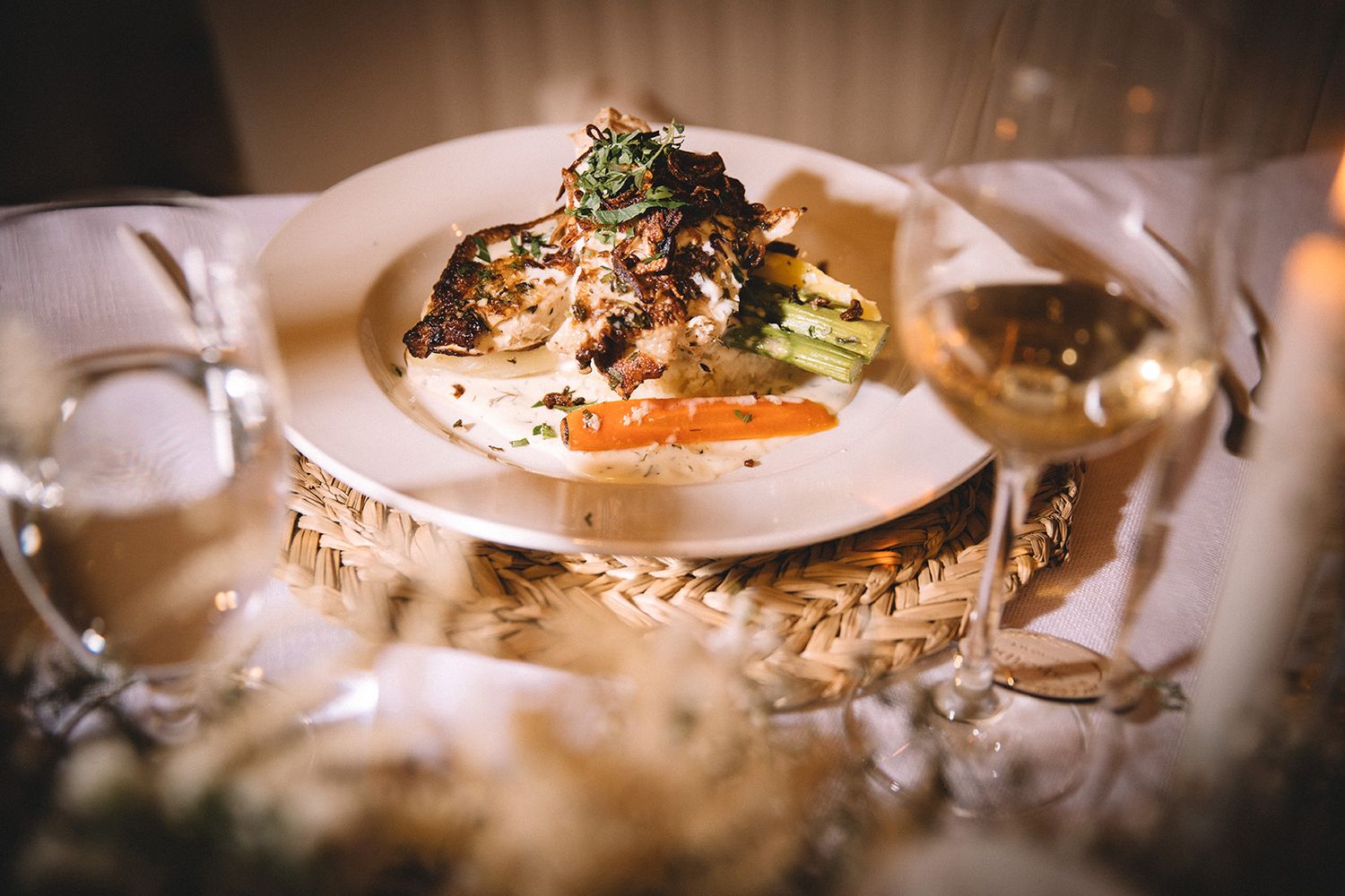 Elegant plated dinner with herbs and white wine glass on textured placemat in romantic lighting.