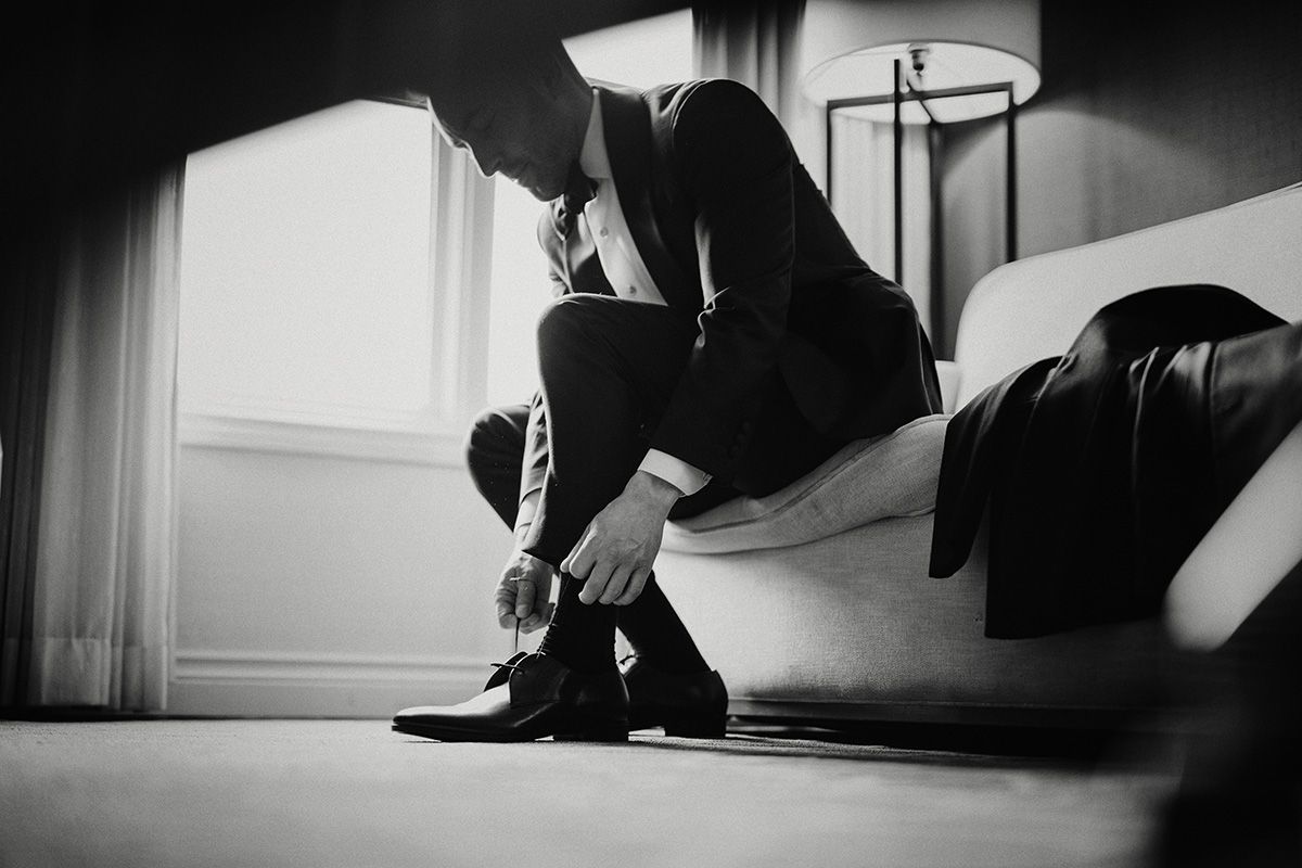 Black and white photo of a groom in a dark suit bending over to tie his shoes.