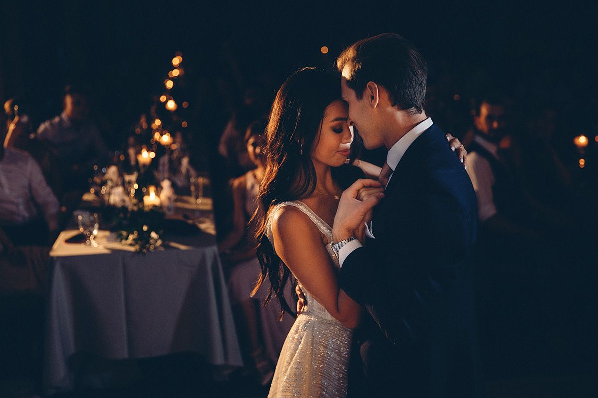 Romantic couple shares an intimate dance together at an elegant wedding reception with candlelit tables in background.
