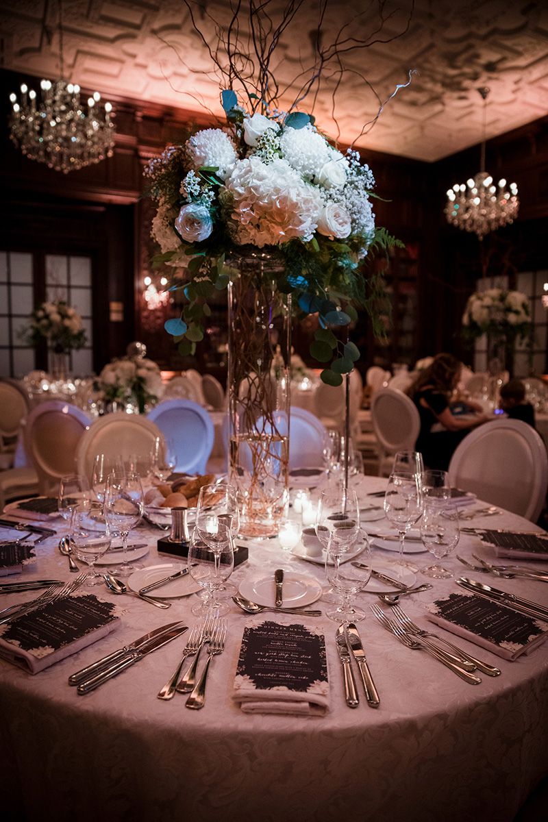 Elegant wedding reception tables with white linens, crystal glassware and tall floral centrepieces in soft blue lighting.