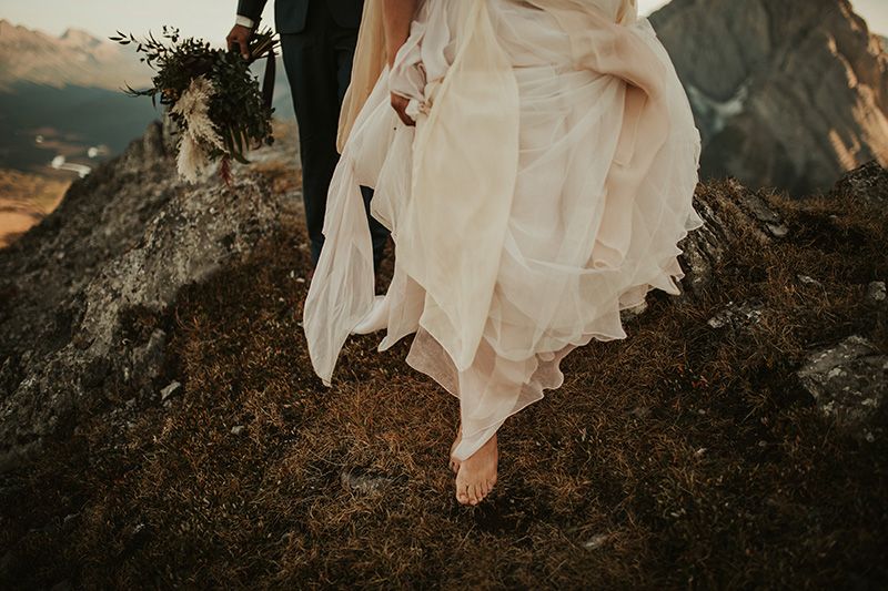 A bride steps barefoot over dark rocks as the wind tosses her white wedding dress creating tails of motion blur.