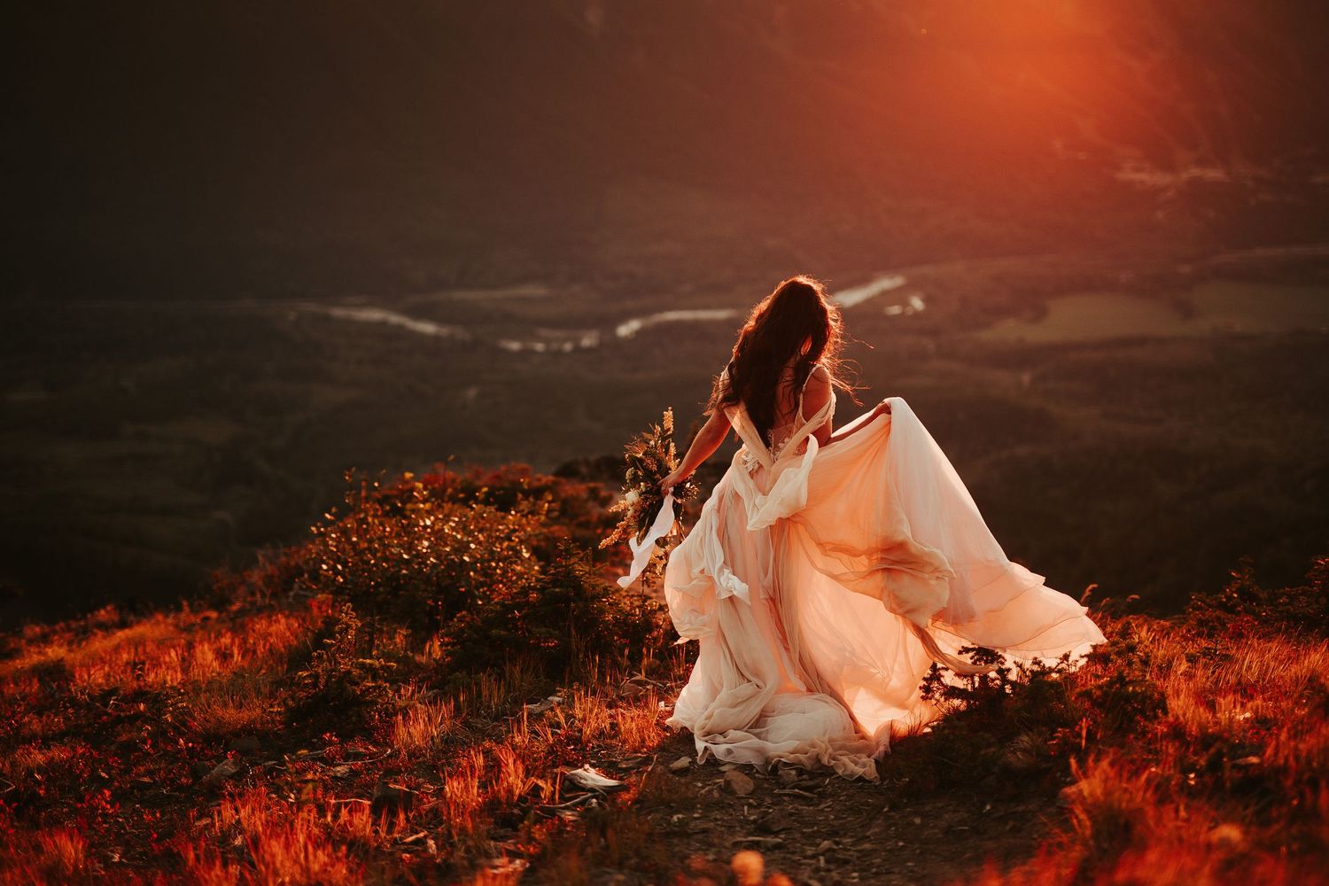 A flowing white dress billows dramatically against a moody red sunset landscape during an artistic outdoor photo session.