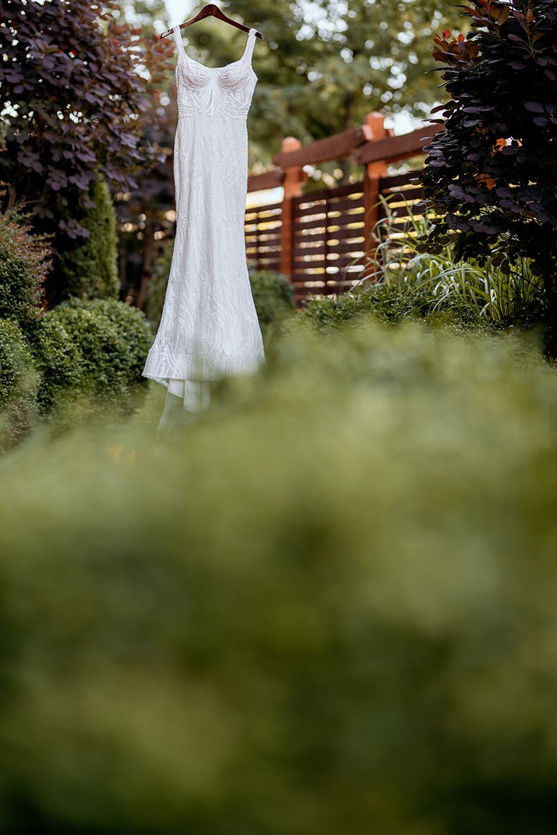 A white wedding dress hanging on a wooden fence in a garden with blurred green foliage in the foreground.
