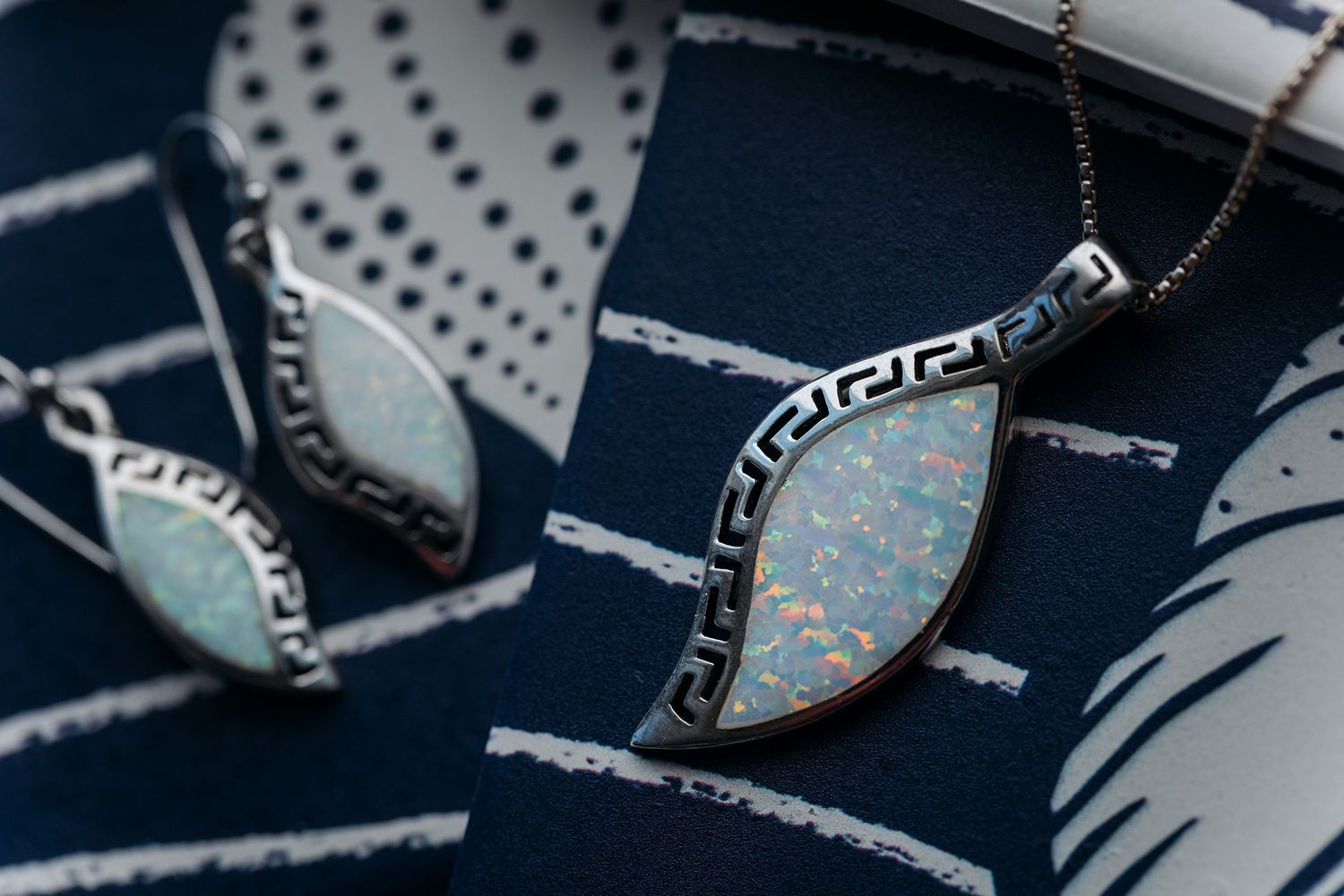 Close-up of elegant silver pendant necklaces with Greek key pattern and light blue stone inlays on dark fabric.