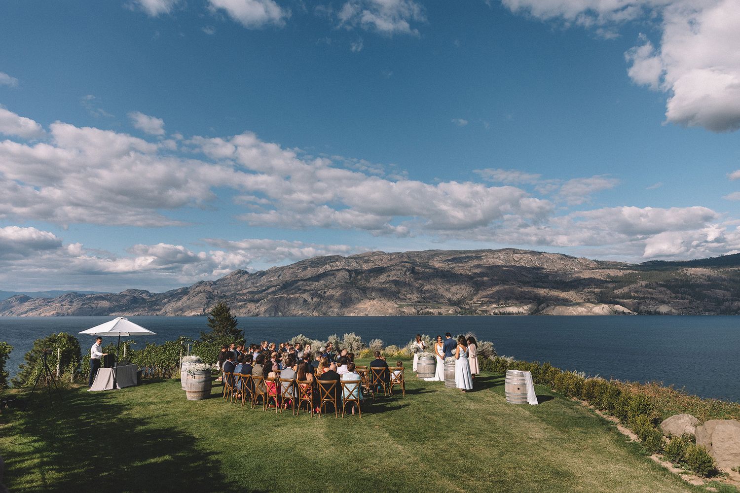 Outdoor wedding ceremony overlooking scenic mountain lake with white chairs and decorations on grassy cliff edge.
