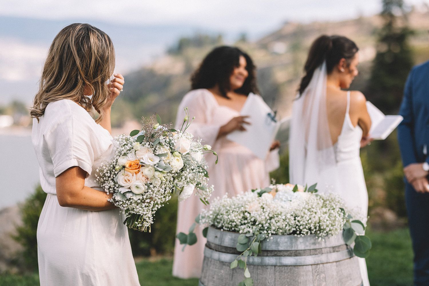 Wedding party members in  pink dresses gather outdoors with floral arrangements displayed on rustic wine barrels.