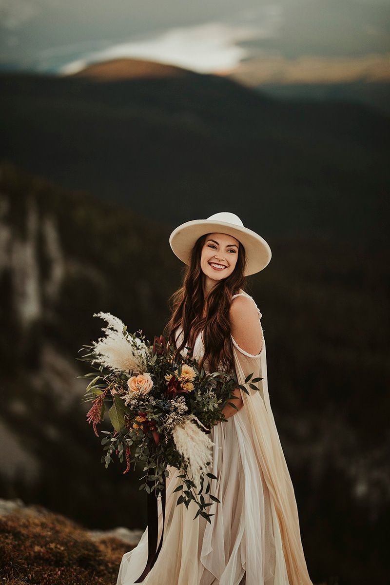 A bride in white dress and sun hat holds bouquet against dramatic mountain landscape at sunset.