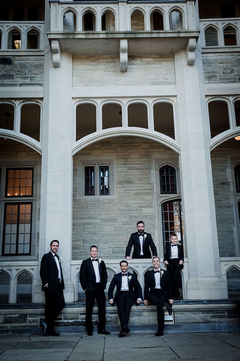 Groomsmen in black tuxedos pose against an elegant stone building facade with arched architecture.