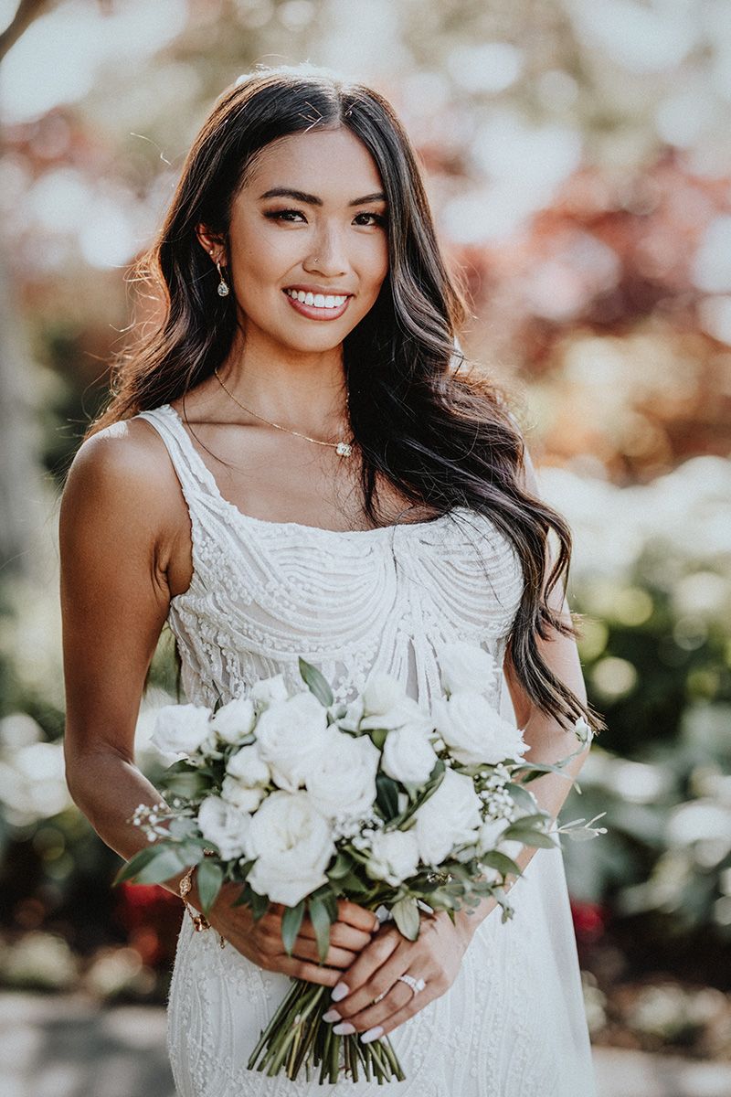 Bride in white lace dress holds elegant white floral bouquet against soft blurred background at her Harvest Golf Club wedding.