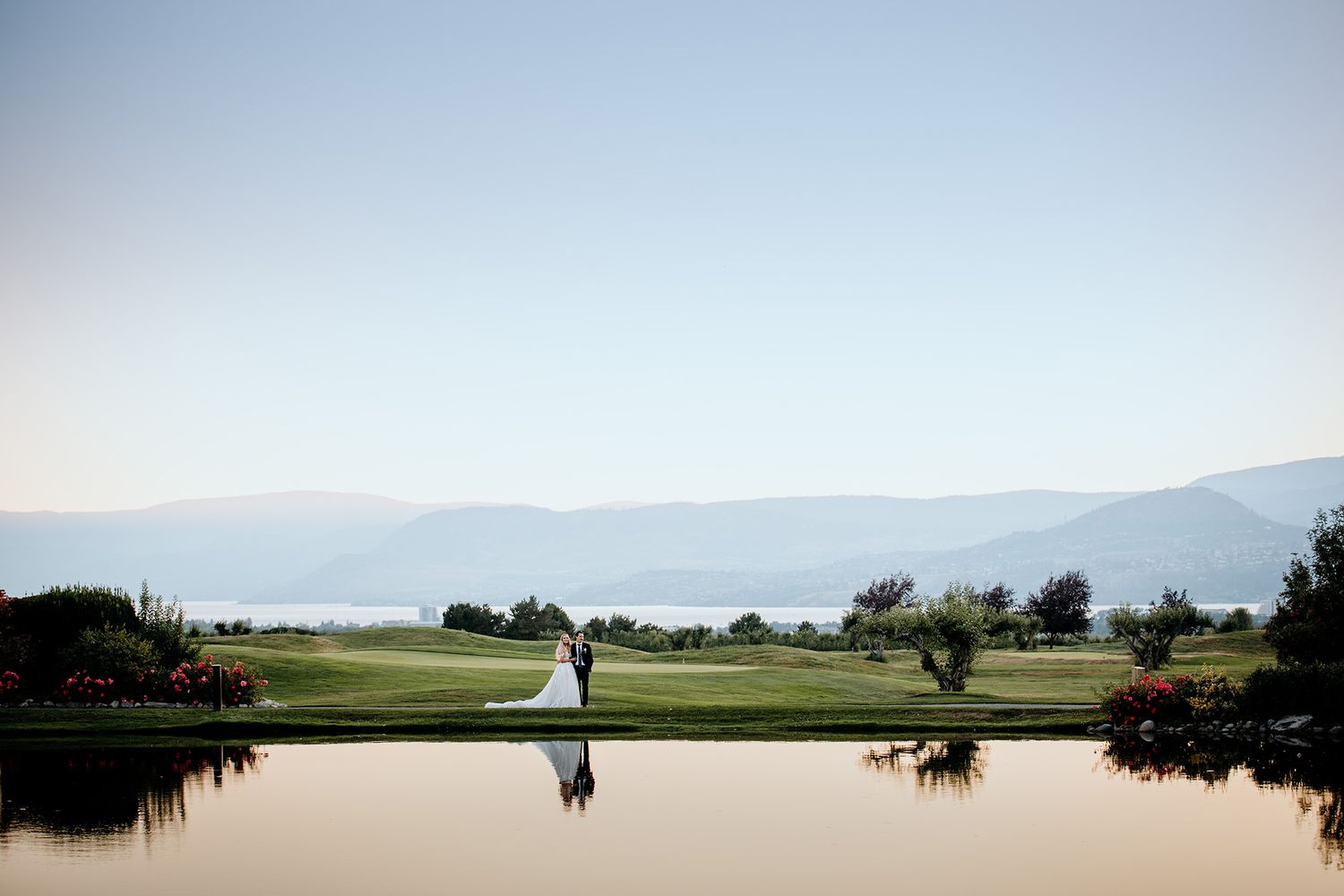 wedding couple walk beside a pond with lake and mountains in the background.