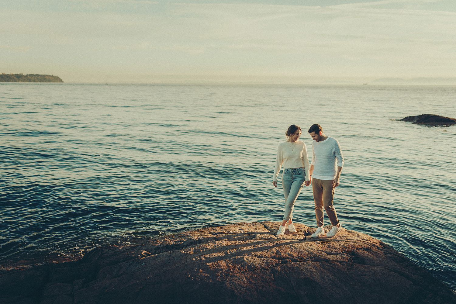Two people stand together on rocky coastal cliffs overlooking a serene ocean at sunset.