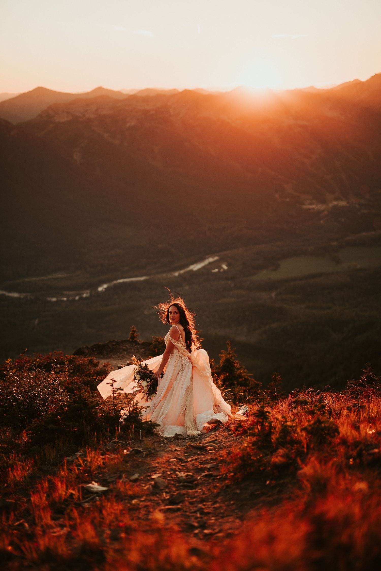 A bride sits atop a mountain ridge at sunset surrounded by vibrant red wildflowers and sweeping valley views.