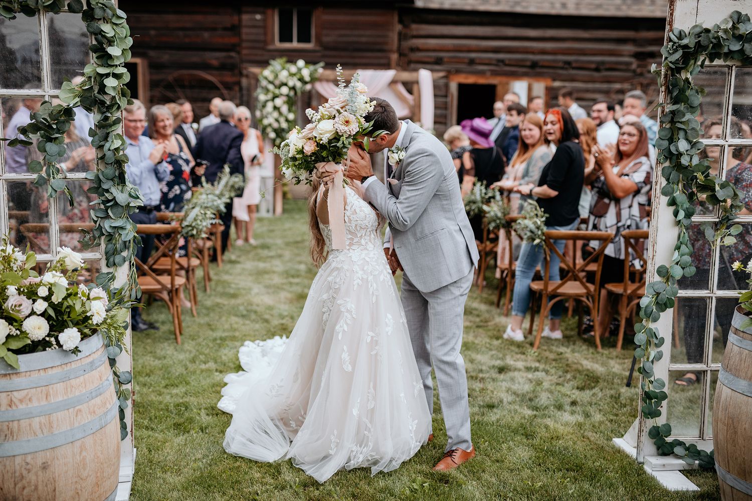 A bride and groom share a kiss during wedding ceremony at a rustic barn venue with wine barrels and floral decorations in the garden.