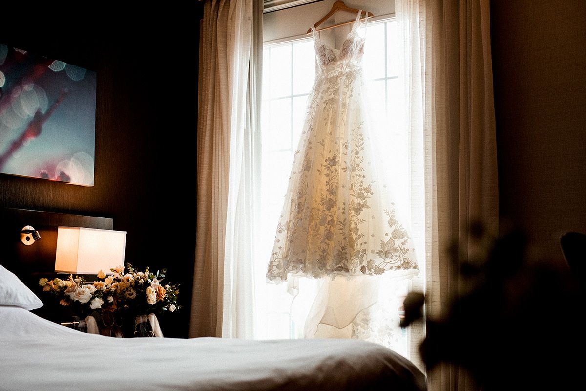 A white lace wedding dress hangs near a sunlit window creating a romantic silhouette against soft natural light.