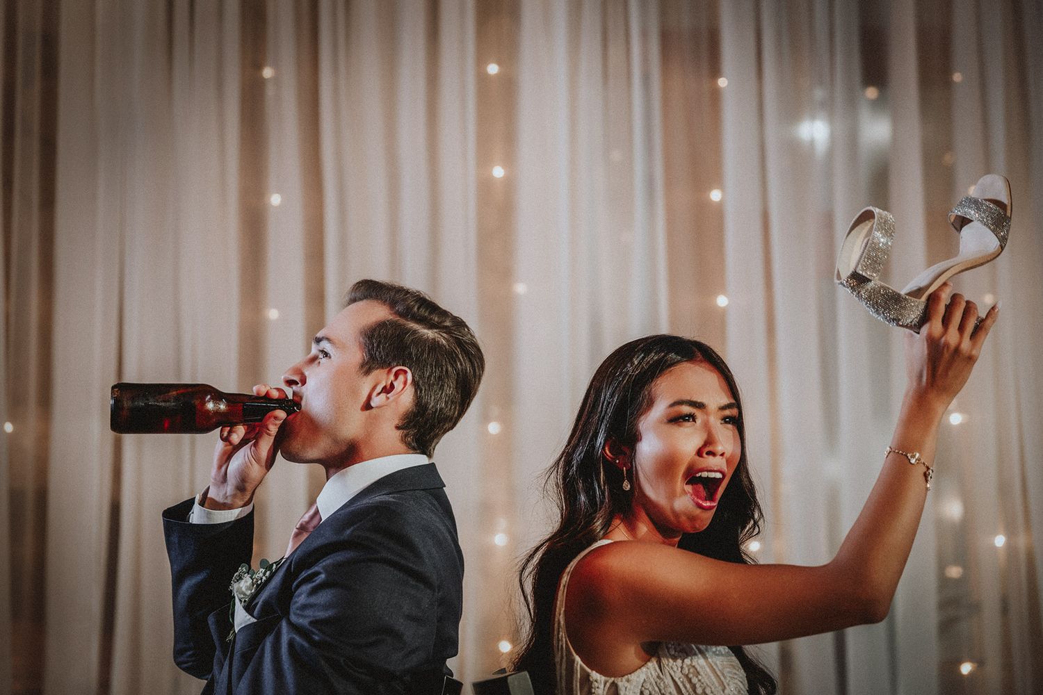 A bride and groom celebrate joyfully at a reception, one drinking from a bottle while the other raises a shoe.