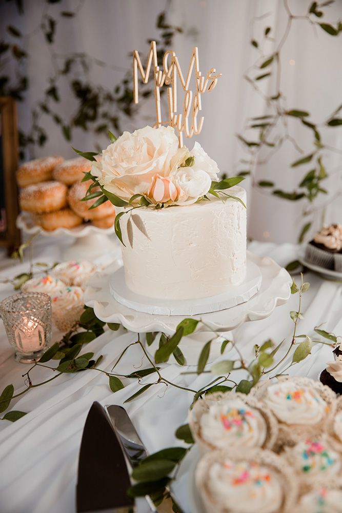 White wedding cake with delicate floral decorations and cupcakes arranged on a table with greenery accents.