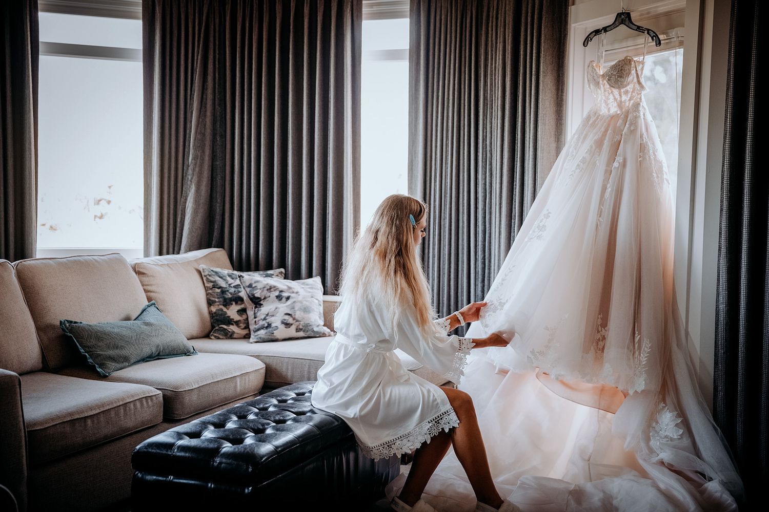 A bride inspects her elegant wedding dress hanging gracefully in a modern hotel room with dark curtains.