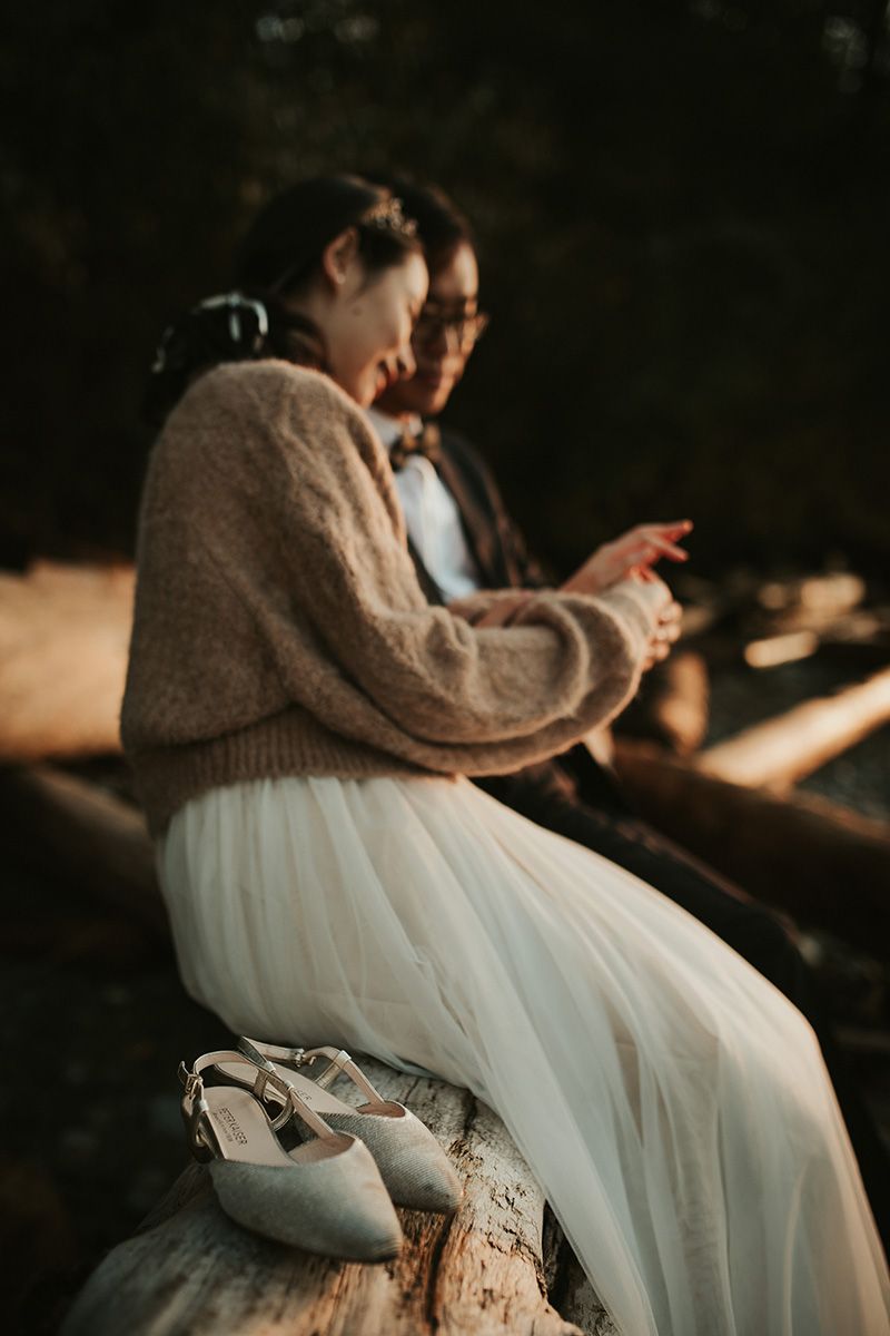 A couple sits together on a rustic bench wearing a white tulle dress and brown knit sweater in a romantic outdoor setting.