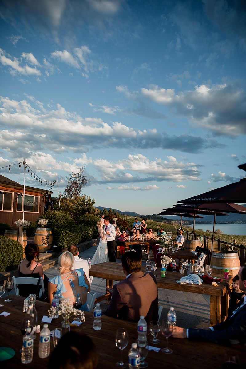 A wedding couple enjoy their first dance at an outdoor reception at sunset with mountain and lake views with cloudy blue skies overhead.