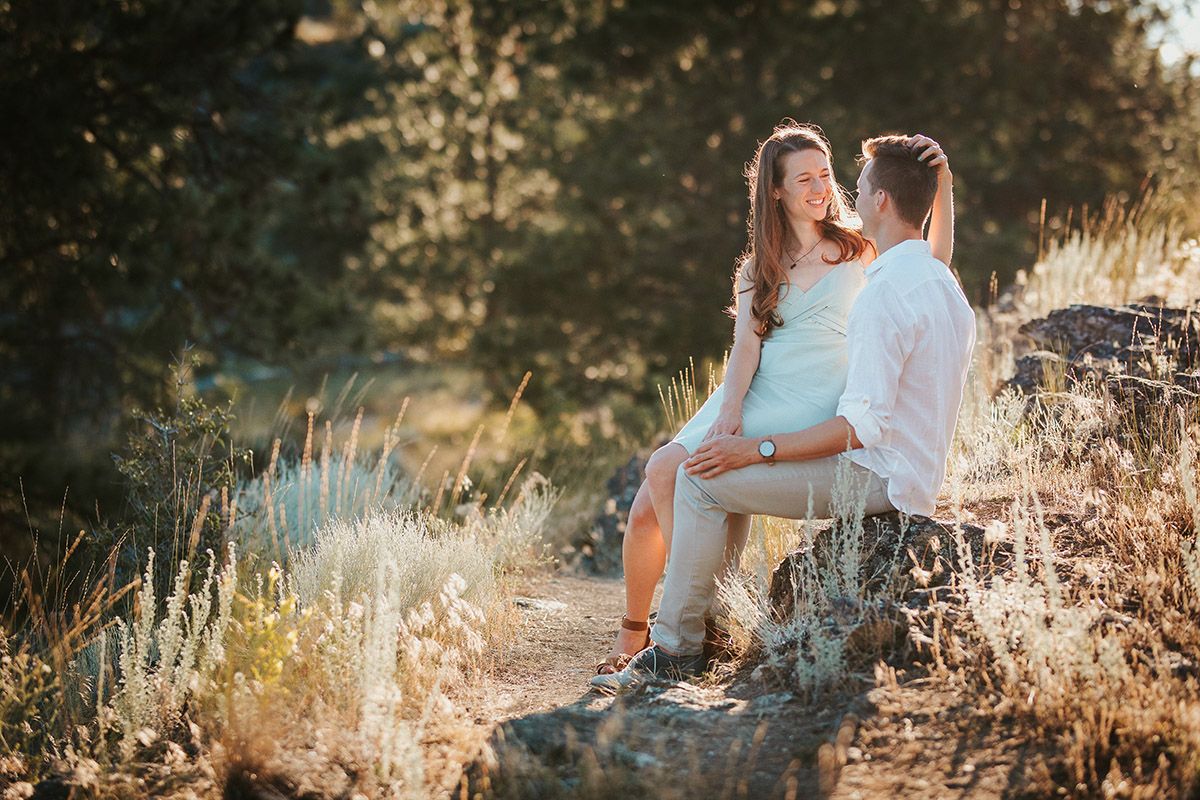 A couple sits on rocks in a sunlit forest, sharing a tender moment surrounded by golden wildflowers.