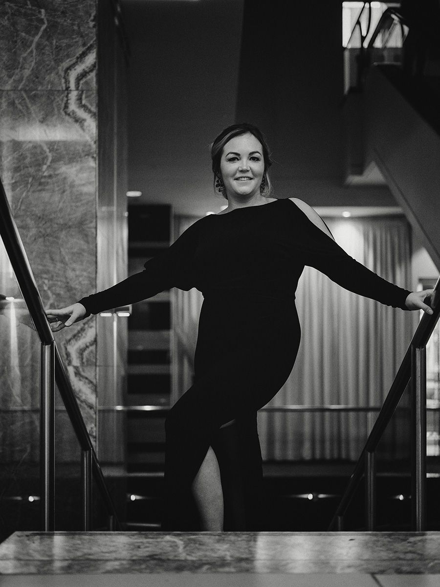 A woman in a black dress strikes an elegant pose against a dark background in this dramatic black and white photograph.
