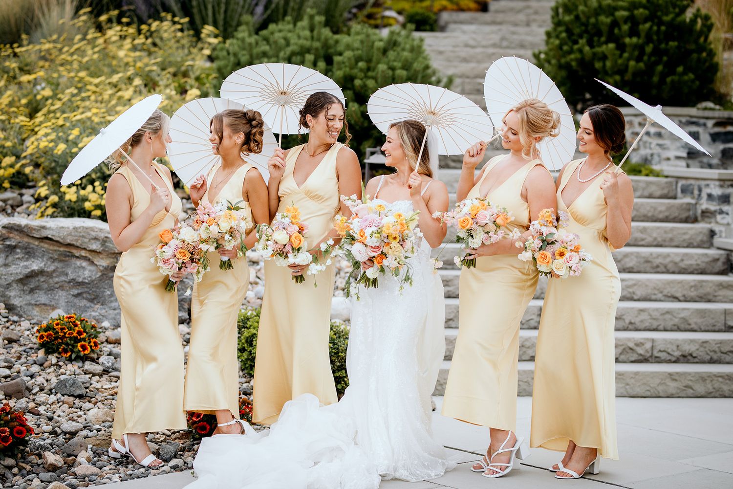 A bridal party in pale yellow dresses, parasols and white gowns stand together on stone steps during an outdoor wedding celebration.