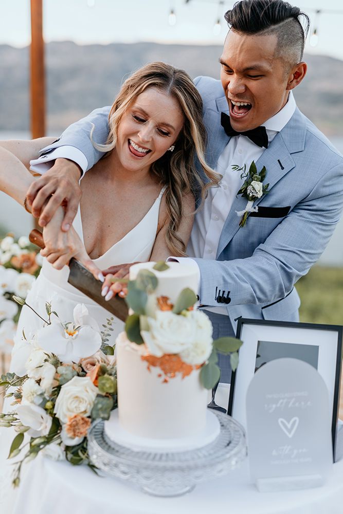 A couple shares joyful moments while cutting their elegant white wedding cake at an outdoor mountain venue.