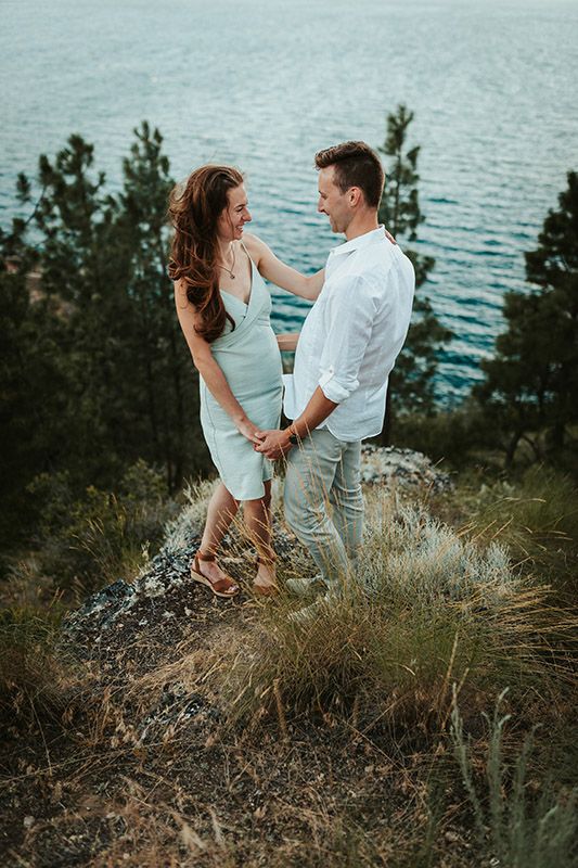 Couple holding hands on a rocky hillside overlooking turquoise water, surrounded by pine trees and wild grass.