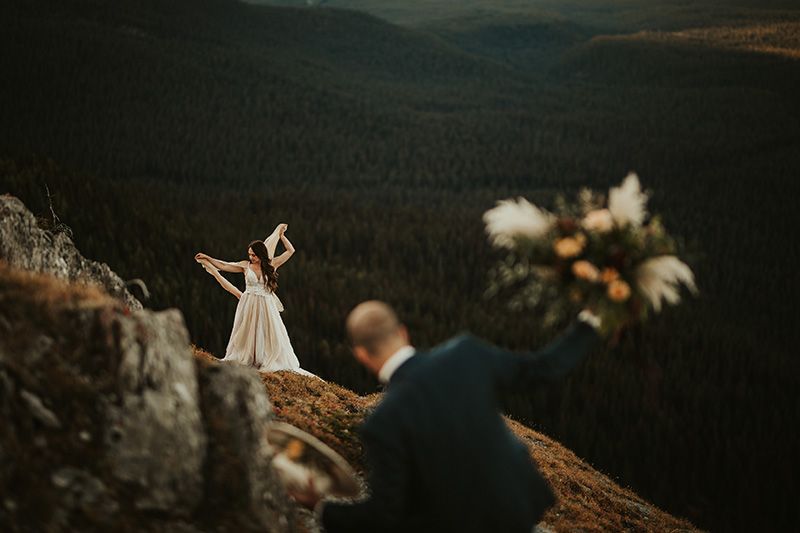 A couple shares an intimate wedding moment on a rocky mountain overlook with a vast forested valley below.