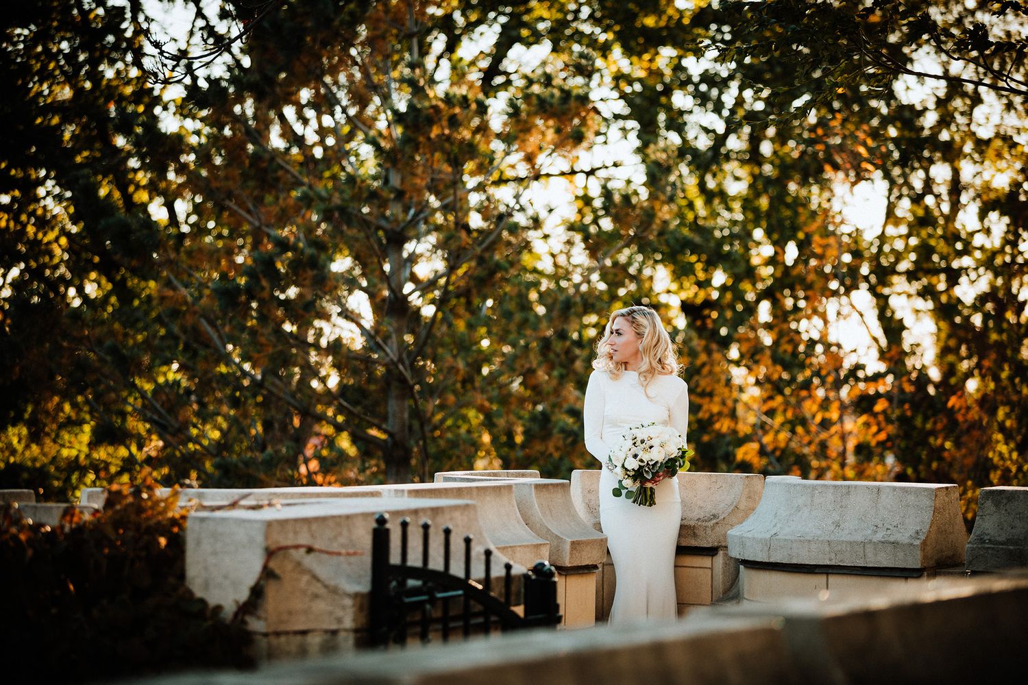 Bride in a white dress holding a bouquet stands on stone steps during golden hour with sunlit trees in background.