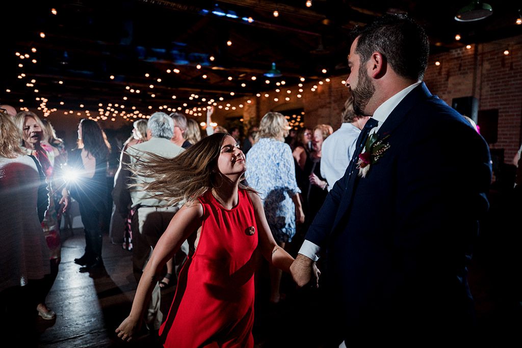 A group of people dancing and celebrating at a wedding reception with string lights overhead in a rustic venue.