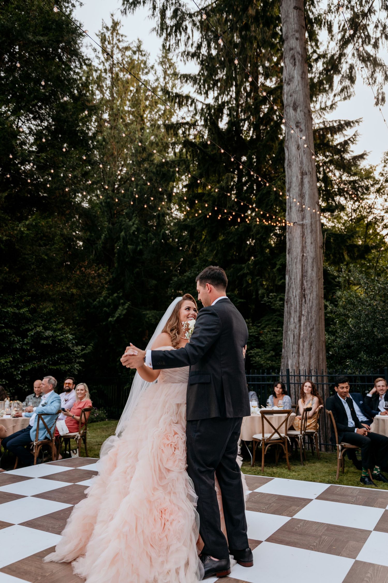 A couple shares a romantic dance on an outdoor checkered dance floor surrounded by tall trees at dusk.