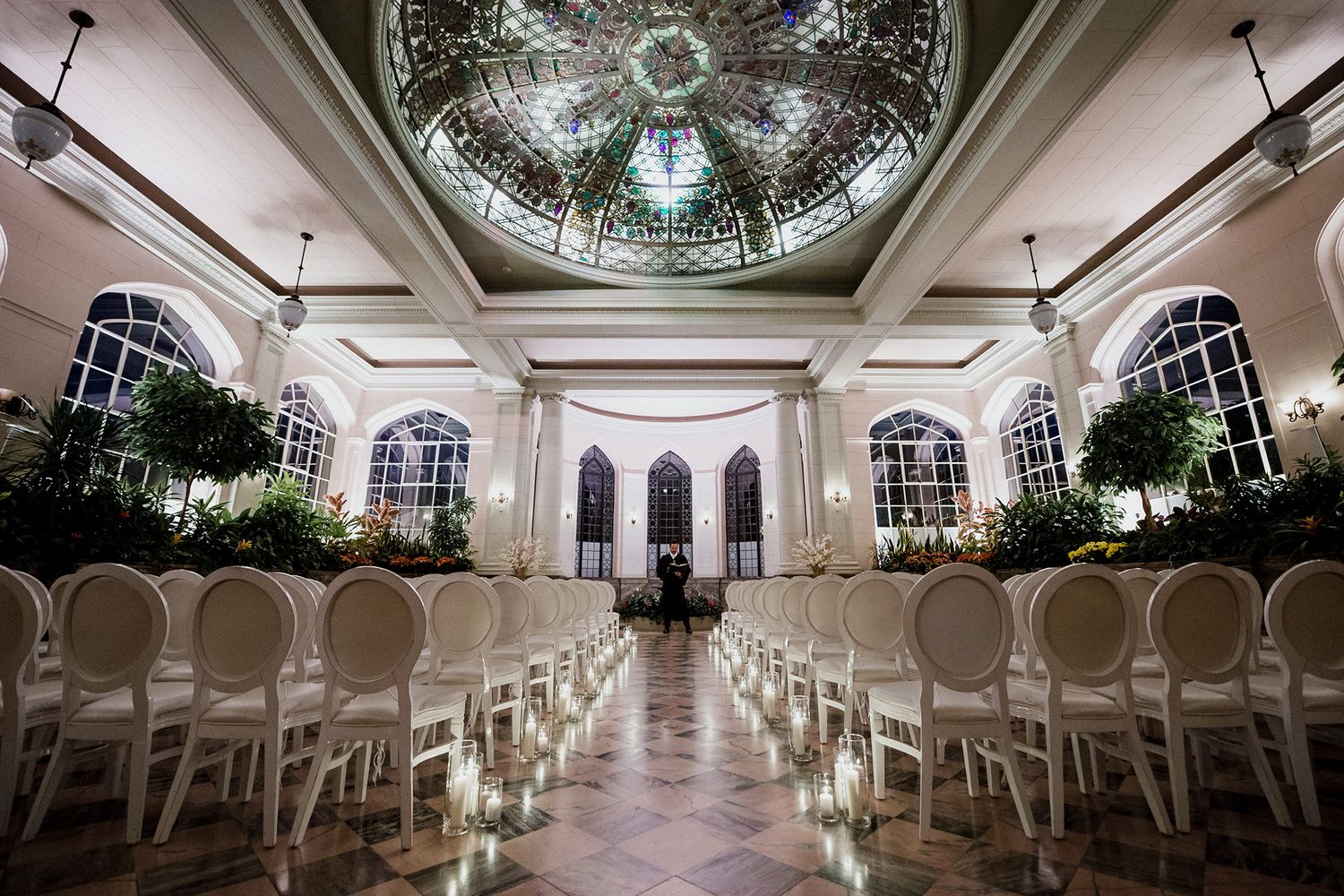Elegant wedding venue interior with white chairs and ornate domed ceiling featuring crystal chandeliers and mirrored details.